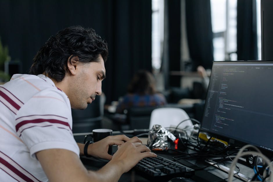 A young man with dark, curly hair is seated at a desk in an office environment, focusing on a computer monitor displaying lines of code. The workspace includes a black keyboard, a black mug, and scatt